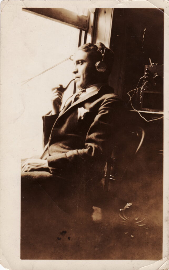 "Papa," a man who was 29 years old in 1924, dressed in a suit. He wears headphones and smokes a pipe. Behind him is an old-fashioned, handmade radio. 